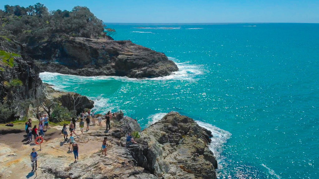 fotografia aérea de la isla Stradbroke de Australia en la que se ve a unos estudiantes encima de las rocas mirando al mar como las olas golpean las rocas